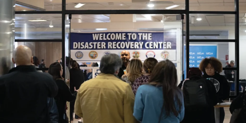 A photo of a crowd inside the West LA Disaster Recovery Center. A man speaks to the crowd with a "Welcome to the Discovery Recovery Center" banner in the background. A UCLA sign is also in the background.