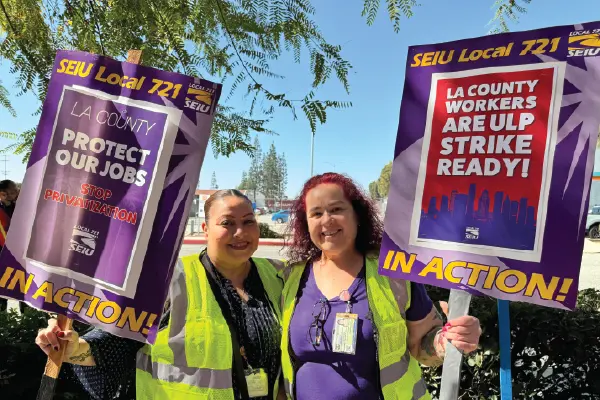 Two union members wearing yellow vests and holding picket signs march outside the Telstar/Aerojet building in El Monte.