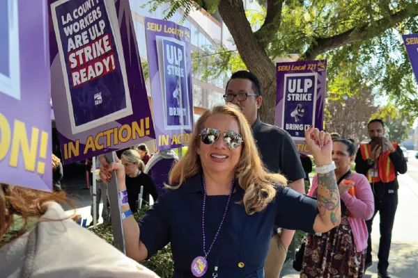A smiling woman in aviator shades holding a picket sign marches with her fellow union members outside the Telstar/Aerojet building in El Monte.