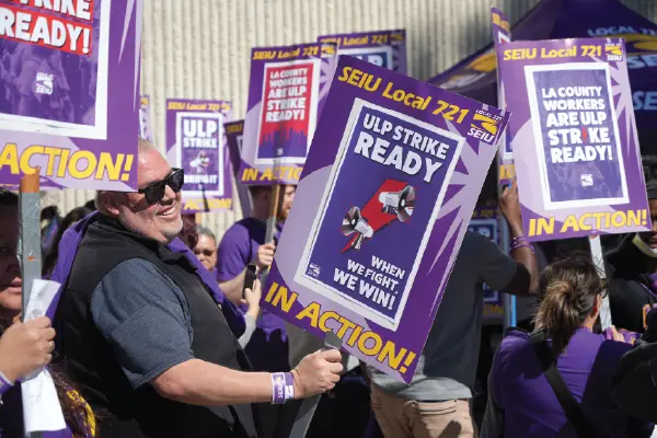 A smiling union member wearing dark sunglasses marches with his fellow union members holding picket signs outside the Olive View-UCLA Medical Center in Sylmar.