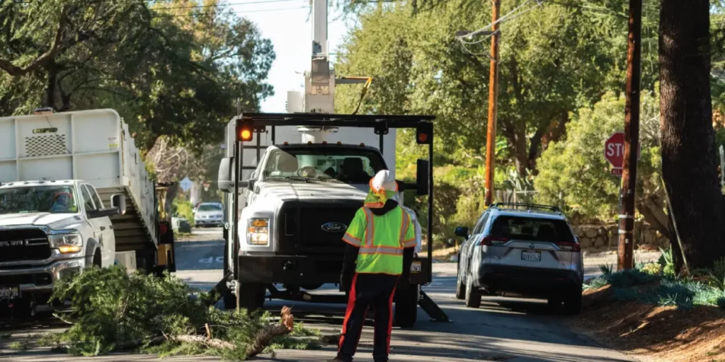 Man wearing a vest in the middle of the street. LA County Public Works truck also in the middle of the street as tree trimming takes place.
