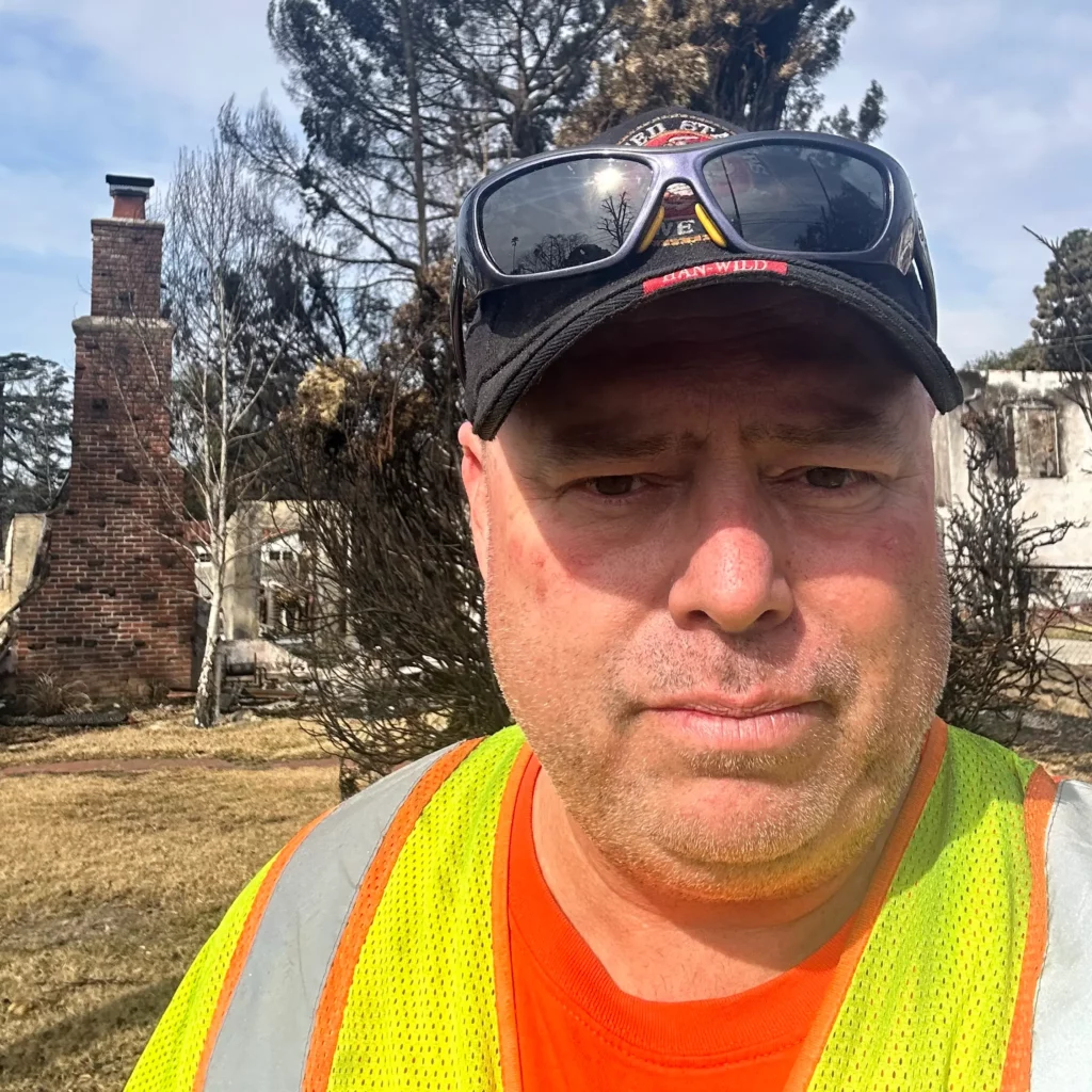 Man with hat and safety vest standing in front of burned down residence. There's also trees in the background.