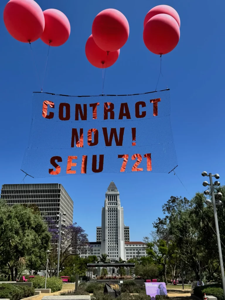 A massive, balloon-held banner spells out "CONTRACT NOW! SEIU 721" in the middle of Grand Park directly outside the LA County Hall of Administration.