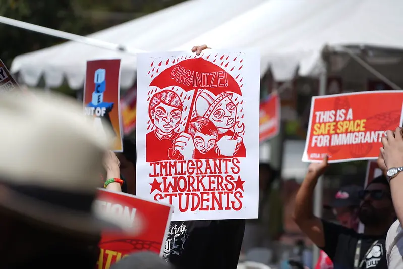 A sign depicts three workers underneath an umbrella with the words "Organize" on it and the slogan "Immigrants, Workers, Students" below it.